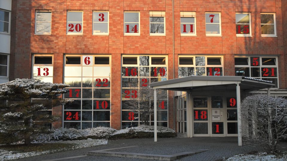 Das Bild zeigt den Eingangsbereich des Polizeipräsidiums Bielefeld bei winterlichem Wetter. Auf den großen fensterelementen sind die Zahlen der Türchen für den Adventskalender in roter Schriftfarbe zu sehen.