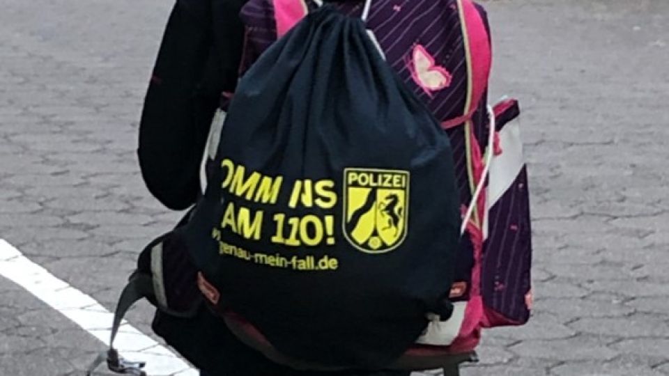 Auf dem Bild ist ein Schulkind mit Schulranzen und Stoffbeutel, mit der Aufschrift "Komm ins Team 110", auf dem Rücken von hinten zu sehen. 