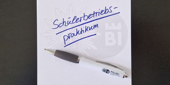 Das Bild zeigt einen Schreibblock mit Wasserzeichen der Polizei Bielefeld, auf dem Blatt steht diagonal "Schülerbetriebsprakikum" geschrieben. Unter dem Schriftzug liegt eine Kugelschreiber mit einem aufgedruckten Logo der Polizei Bielefeld.